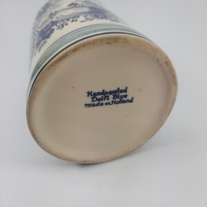 May include: A hand-painted Delft Blue ceramic mug. The mug has a blue and white landscape design. The base of the mug is inscribed with "Handpainted Delft Blue made in Holland".