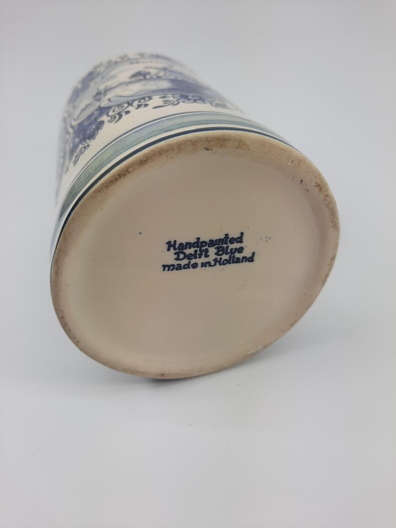 May include: A hand-painted Delft Blue ceramic mug. The mug has a blue and white landscape design. The base of the mug is inscribed with "Handpainted Delft Blue made in Holland".