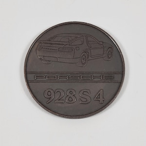 Puede incluir: Una moneda redonda de metal gris oscuro con un contorno en blanco y negro de un coche Porsche 928 S4. La moneda tiene las palabras "PORSCHE" y "928 S 4" grabadas.