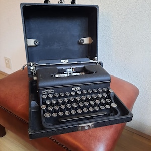 May include: A black Royal typewriter in its open carrying case. The typewriter has a full keyboard with silver keys and a black carriage. The case is open, revealing the typewriter inside. The case has a black handle.