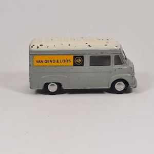 May include: A vintage gray toy delivery van with a white roof. The van has black wheels and a yellow rectangle with the text "VAN GEND & LOOS" and a logo. A classic collectible toy.
