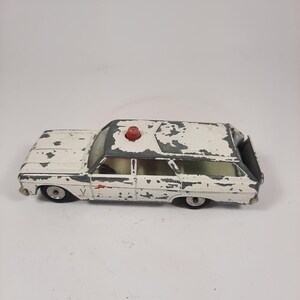 May include: A vintage white and gray toy station wagon with a red light on top. The toy car shows signs of wear, with paint chipping off the body. The wheels are black and the windows are clear.