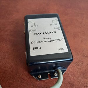 Op de afbeelding: Een zwarte metalen doos met een zilveren label waarop staat "MONACOR Stereo Entzerrvorverstärker (RIAA) SPR-4 JAPAN". De doos heeft twee ingangs- en twee uitgangsconnectoren.