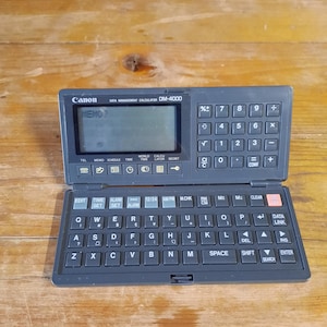 Könnte beinhalten: Ein schwarzer Canon DM-4000 Datenmanagement-Rechner mit einem kleinen LCD-Bildschirm und einer QWERTY-Tastatur. Der Bildschirm zeigt den Text "MEMO?" an.