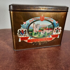 May include: A gold metal tin with a vintage design. The tin features a castle illustration, the words "Marca Selecta" and "Balmoral", and the text "Tabacos Exquisitos".