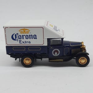 May include: A miniature vintage-style delivery truck featuring a dark blue cab and a white cargo box. The side of the cargo box displays the Corona Extra logo in blue and gold. The truck has gold-colored wheels and a circular emblem on the door.