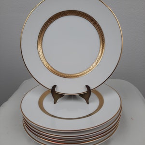 Puede incluir: Un conjunto de seis platos llanos blancos con un borde dorado y un dise&ntilde;o dorado. Los platos est&aacute;n apilados sobre una superficie blanca.