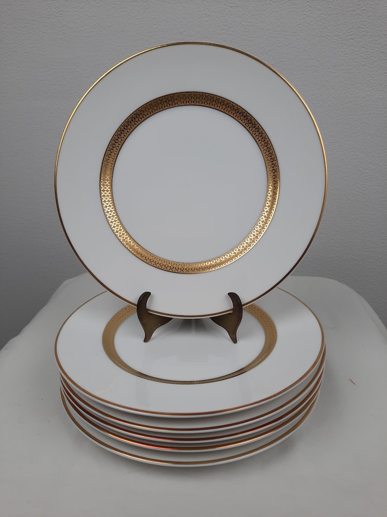Puede incluir: Un conjunto de seis platos llanos blancos con un borde dorado y un dise&ntilde;o dorado. Los platos est&aacute;n apilados sobre una superficie blanca.
