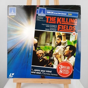 Puede incluir: Una portada de disco de vinilo azul y rojo para la banda sonora de la película "The Killing Fields". La portada presenta una foto de una escena de la película con tres hombres que sostienen armas de fuego. El texto de la portada dice "The Killing Fields" y "Música original compuesta por Mike Oldfield".