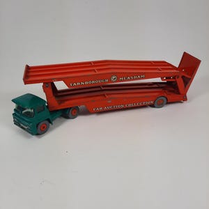 Puede incluir: Un camión de juguete transportador de coches vintage en rojo y verde. El camión tiene las palabras "FARNBOROUGH MEASHAM" y "CAR AUCTION COLLECTION" impresas. El camión tiene una rampa que se puede bajar.