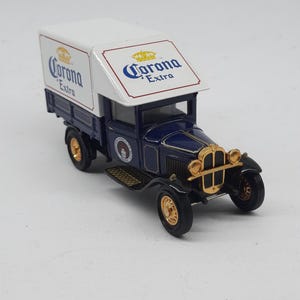 May include: A die-cast model truck featuring a dark blue cab and a white cargo box with Corona Extra branding. The vehicle has gold-colored accents on the front grille, headlights, and wheels, with a vintage design and detailed side logos.