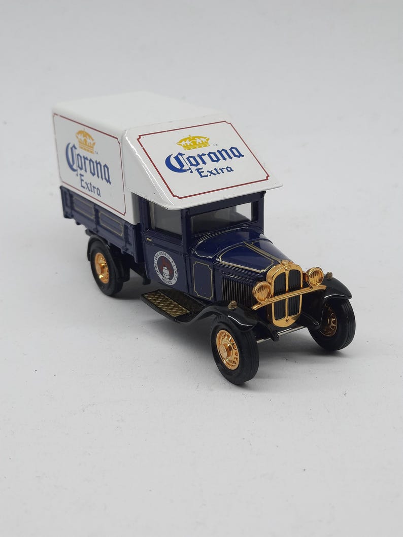 May include: A die-cast model truck featuring a dark blue cab and a white cargo box with Corona Extra branding. The vehicle has gold-colored accents on the front grille, headlights, and wheels, with a vintage design and detailed side logos.