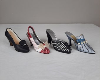 May include: Three miniature high-heeled shoes. The first shoe is black with a pointed toe. The second shoe is white with red polka dots and a red bow. The third shoe is light blue with silver embellishments.