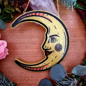 Pode incluir: Um pendurado de parede de lua crescente dourado com um rosto e l&aacute;bios vermelhos. A lua &eacute; decorada com glitter dourado e tem um contorno preto. A lua est&aacute; pendurada em uma corrente dourada.