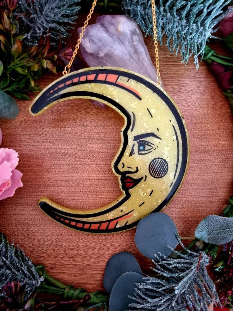 Pode incluir: Um pendurado de parede de lua crescente dourado com um rosto e l&aacute;bios vermelhos. A lua &eacute; decorada com glitter dourado e tem um contorno preto. A lua est&aacute; pendurada em uma corrente dourada.