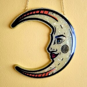 Pode incluir: Um pendurado de parede em forma de lua crescente com um rosto e glitter embutido na resina. A lua &eacute; amarela com contornos pretos e detalhes vermelhos. O rosto tem um olho azul e uma boca vermelha.