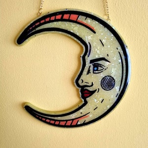 Pode incluir: Um ornamento de resina em forma de lua crescente com acabamento brilhante. A lua tem um rosto com um olho azul, l&aacute;bios vermelhos e um c&iacute;rculo preto e branco na bochecha. O ornamento est&aacute; pendurado em uma corrente dourada.