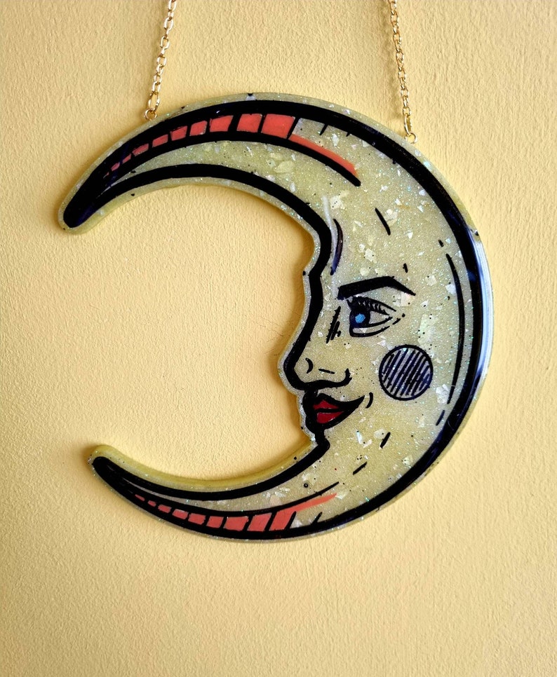 Pode incluir: Um ornamento de resina em forma de lua crescente com acabamento brilhante. A lua tem um rosto com um olho azul, l&aacute;bios vermelhos e um c&iacute;rculo preto e branco na bochecha. O ornamento est&aacute; pendurado em uma corrente dourada.