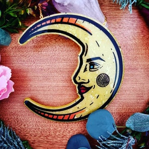 Pode incluir: Uma lua crescente dourada com um rosto pintado nela. A lua &eacute; feita de resina e tem um acabamento brilhante.