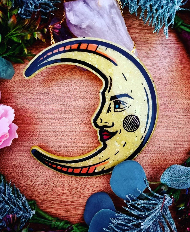 Pode incluir: Uma lua crescente dourada com um rosto pintado nela. A lua &eacute; feita de resina e tem um acabamento brilhante.