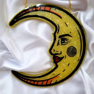 Pode incluir: Uma lua crescente amarela com um rosto, delineado em preto, com uma corrente dourada anexada. A lua tem uma textura brilhante e acentos vermelhos e laranja.
