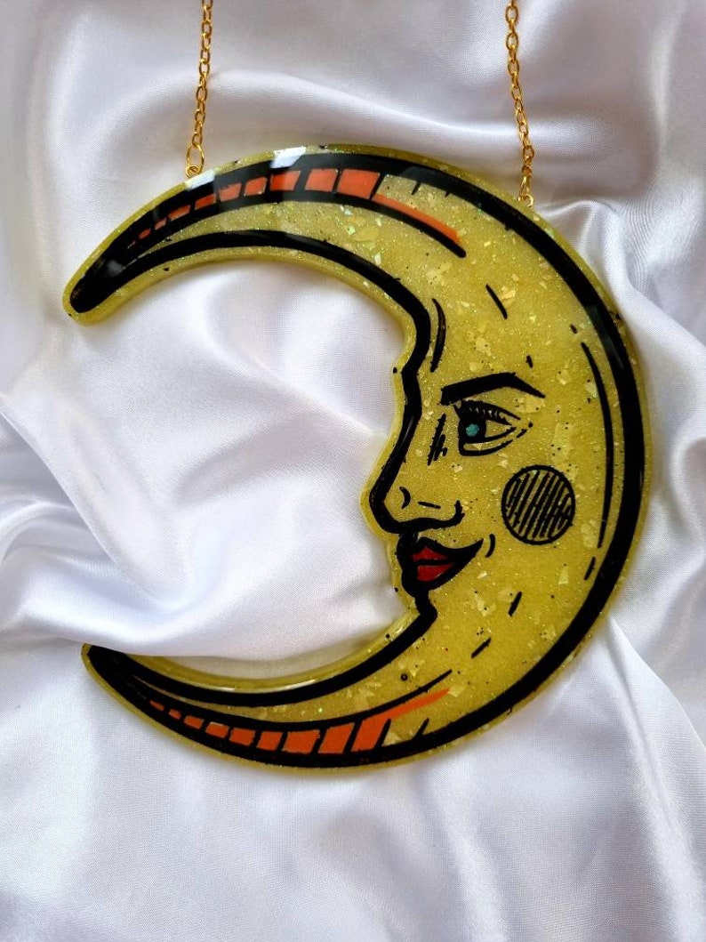 Pode incluir: Uma lua crescente amarela com um rosto, delineado em preto, com uma corrente dourada anexada. A lua tem uma textura brilhante e acentos vermelhos e laranja.