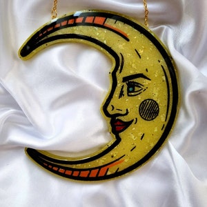 Pode incluir: Uma lua crescente amarela com um rosto desenhado nela. A lua &eacute; feita de resina e tem um acabamento brilhante. Est&aacute; pendurada em uma corrente dourada.