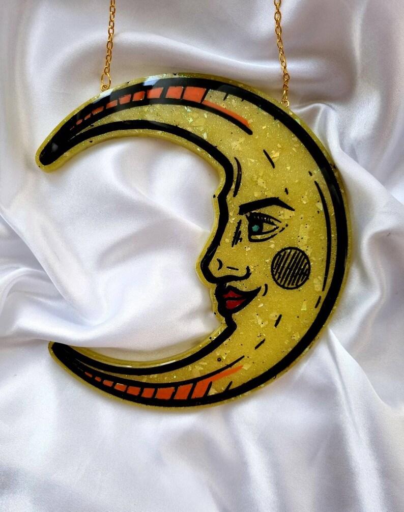 Pode incluir: Uma lua crescente amarela com um rosto desenhado nela. A lua &eacute; feita de resina e tem um acabamento brilhante. Est&aacute; pendurada em uma corrente dourada.