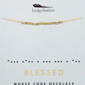 May include: Gold necklace with small beads and bars spelling out "BLESSED" in Morse code. The necklace is on a light blue background with a gold grid pattern. The text "MORSE CODE NECKLACE" is below the necklace.