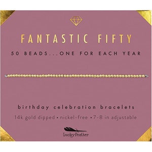 May include: A gold bracelet with 50 beads, one for each year. The card reads "Fantastic Fifty 50 Beads...One for Each Year" and "Birthday Celebration Bracelets 14k gold dipped nickel-free 7-8 in adjustable". The card is pink with gold accents and a diamond shape in the top corner. The brand name "Lucky Feather" is printed in black on a white feather.