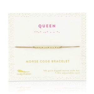 May include: A Morse code bracelet on a white card. The card has the word "QUEEN" in pink and the Morse code equivalent. The bracelet has a gold-colored bar with dots and dashes, strung on a tan cord. The card also says "MORSE CODE BRACELET".