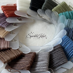 Puede incluir: Una disposición circular de madejas de hilo de bordar enrolladas en bobinas individuales de plástico translúcido en una variedad de colores que incluyen óxido, blanco, negro, verde, azul, marrón y gris, con el texto Sparkle Knot STUDIO en el centro.