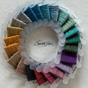 May include: A circular collection of metallic embroidery thread samples wound onto individual translucent plastic bobbins, arranged in a color gradient from blue and green to purple, red, black, and gold, with the text Sparkle Knot STUDIO in the center.
