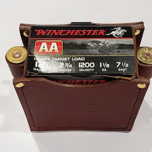 May include: A brown leather shotgun shell holder with gold rivets, holding a box of Winchester AA 12 gauge shells. The box has red and black text, and the holder has two side pockets with shells. The text "DEAD BIRD PRODUCTS" is visible.