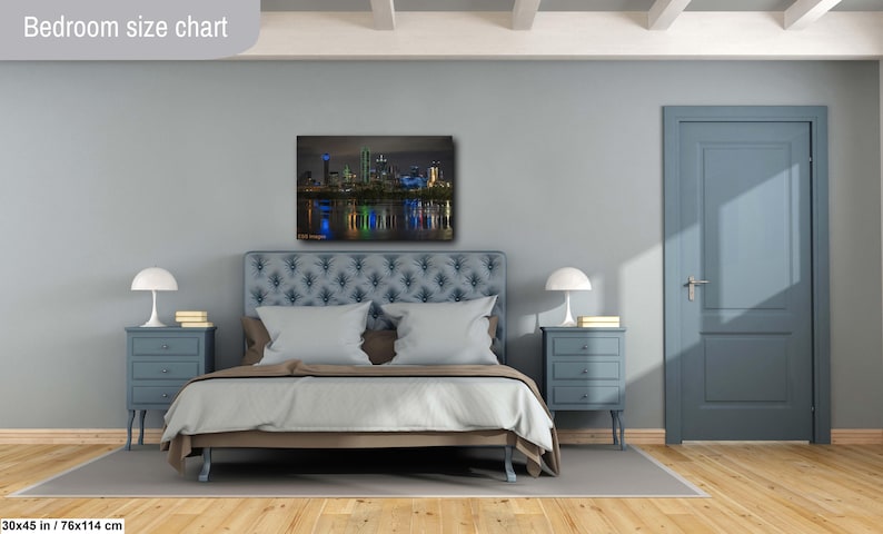 May include: A bedroom with a blue wall and a bed with a blue headboard. A cityscape canvas print hangs above the bed. The print shows a city skyline at night with lights reflecting in the water. Two nightstands with lamps are on either side of the bed.