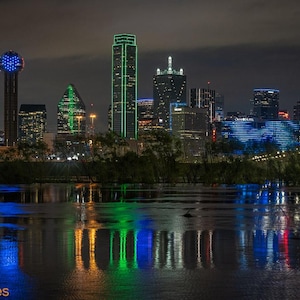 May include: A night-time view of the Dallas skyline, featuring several skyscrapers lit up in blue and green. The buildings are reflected in the water below.