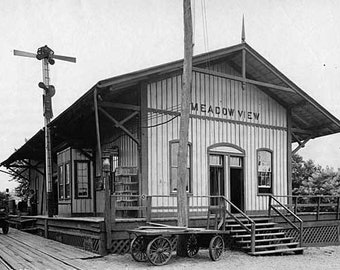 May include: A black and white photograph of a train station with the sign "MEADOW VIEW" above the entrance. The station is made of wood and has a large awning over the platform. There are two people standing on the platform, and a handcart is parked in front of the station.