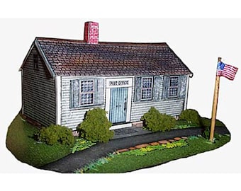 May include: A miniature model of a white post office building with a red brick chimney and a sign that reads "POST OFFICE". The building has a brown roof and two windows on each side. There is a small American flag on a pole in front of the building.
