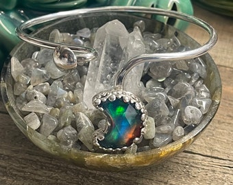 May include: A silver crescent moon charm bracelet with a dark green and iridescent opal stone. The bracelet is resting on a bed of gray stones.