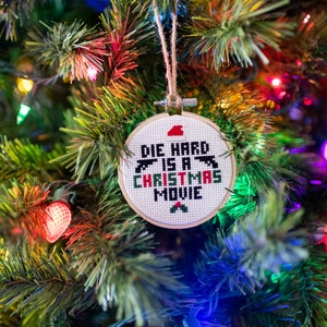 Könnte beinhalten: Ein weißer Weihnachtsbaumschmuck mit roter und grüner Stickerei, auf dem "Die Hard is a Christmas Movie" steht. Der Schmuck hängt an einem grünen Weihnachtsbaum mit bunten Lichtern.