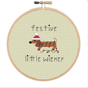 Könnte beinhalten: Ein brauner Dackel mit einem Weihnachtsmann-Hut und Weihnachtslichtern ist auf einem beigefarbenen Stoff gestickt, der über einen hölzernen Stickrahmen gespannt ist. Der Text "festive little wiener" ist unter dem Dackel gestickt.