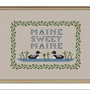 May include: Cross-stitch framed artwork with the words "MAINE SWEET MAINE" in blue. The artwork features two loons in a blue pond, surrounded by a green and brown floral border. The background is a light beige color.
