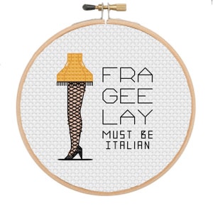 May include: A white embroidery hoop with a yellow lampshade with black legs and black heels. The text "FRA GEE LAY MUST BE ITALIAN" is printed in black.