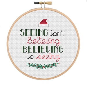 May include: A white embroidery hoop with a green and red Christmas wreath at the bottom. The text "SEEING isn't believing BELIEVING is seeing" is embroidered in black and red thread. A red Santa hat is embroidered at the top.