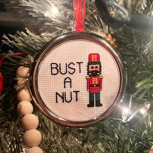 May include: A white Christmas ornament with a silver frame and a red and white embroidered nutcracker. The ornament has the text "Bust a Nut" embroidered on it.