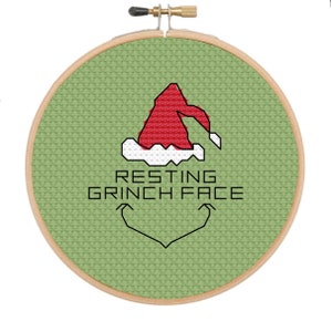 May include: A green embroidery hoop with a cross-stitch design of a Grinch wearing a Santa hat. The text "Resting Grinch Face" is below the design.