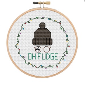 May include: A cross-stitch embroidery hoop with a white background and a green wreath with colorful lights. The wreath surrounds a brown knit hat with a pom-pom and glasses. The text "OH FUDGE" is stitched in green below the hat.