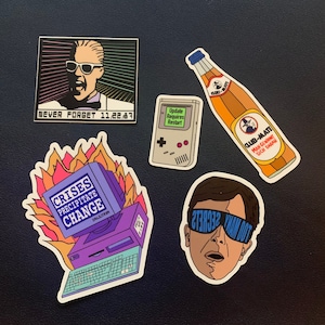 Könnte beinhalten: Fünf bunte Aufkleber mit verschiedenen Designs. Ein Aufkleber zeigt einen Mann mit Sonnenbrille und dem Text "NEVER FORGET 11/22/87". Ein weiterer Aufkleber zeigt eine Retro-Videospielkonsole mit dem Text "Update Requires Restart". Ein dritter Aufkleber zeigt einen Computer mit Flammen und dem Text "CRISES PRECIPITATE CHANGE". Ein vierter Aufkleber zeigt eine Flasche Limonade mit dem Text "CLUB-MATE MAN GEWINNT SICH DARAN". Der letzte Aufkleber zeigt einen Mann mit Sonnenbrille und dem Text "TOO MANY SECRETS".