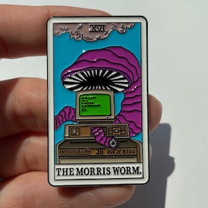 Puede incluir: Un pin de esmalte colorido que representa una computadora con una criatura de gusano púrpura que emerge de la pantalla. El pin es un diseño de carta de tarot con el texto "XVI THE MORRIS WORM." y "INTERGRAPH 36 VAX 8550".