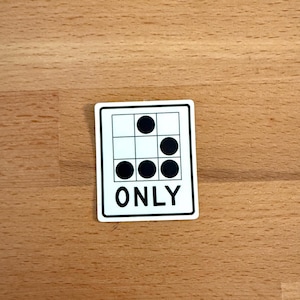 May include: A white square sticker with a black border. The sticker features a grid pattern with black circles in some squares and the word "ONLY" at the bottom. The sticker is on a wooden surface.